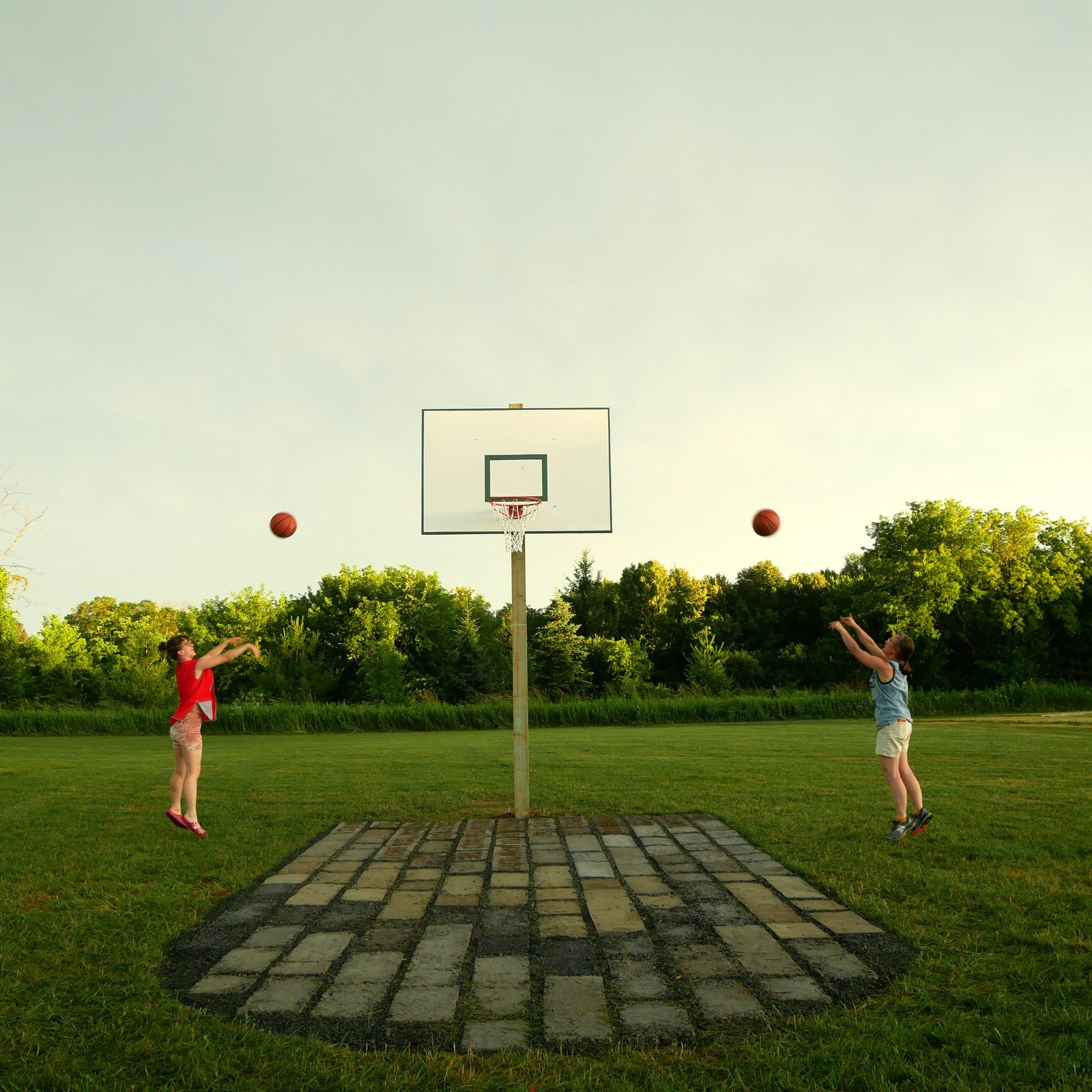 two people shoot basketballs at a hoop against a cloudy sky on a dirtball court 