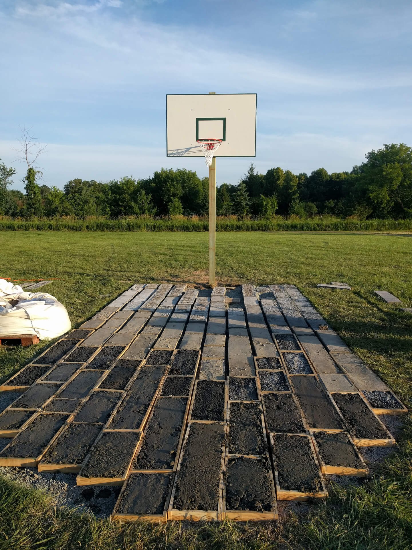 A green and white basketball backboard against a blue sky. Below concrete pavers are still in forms.