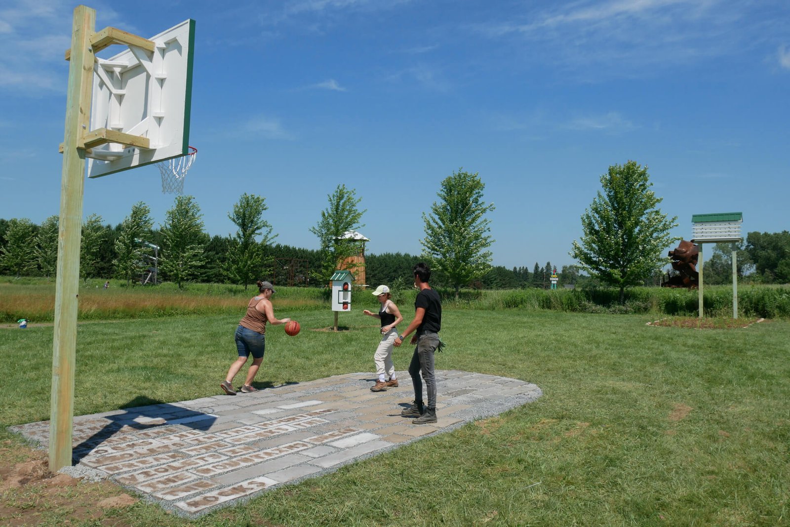 3 people play basketball on a concrete free throw lane that has text carved into it. A small house holds two more basketballs. A garden and a purple martin house.