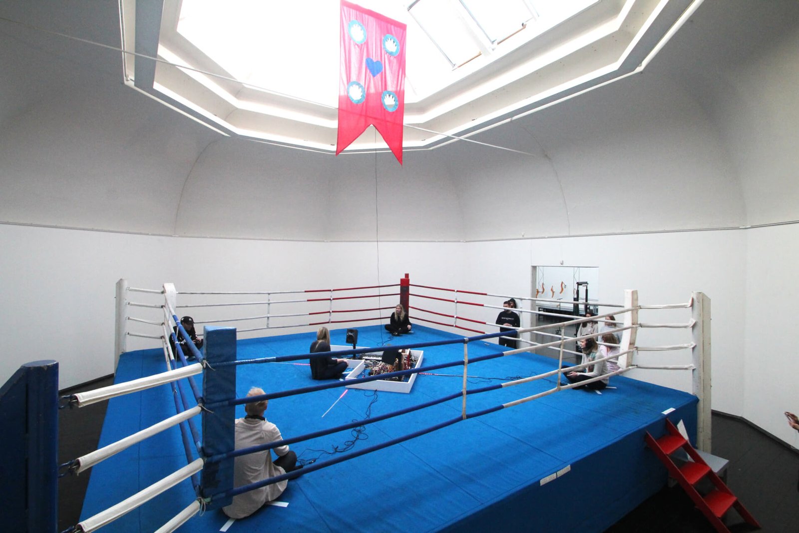 A person in each of a boxing ring's 4 corner's holding a controller that is connected to a instruments in the center where a 5th player's heartbeat drives the music.