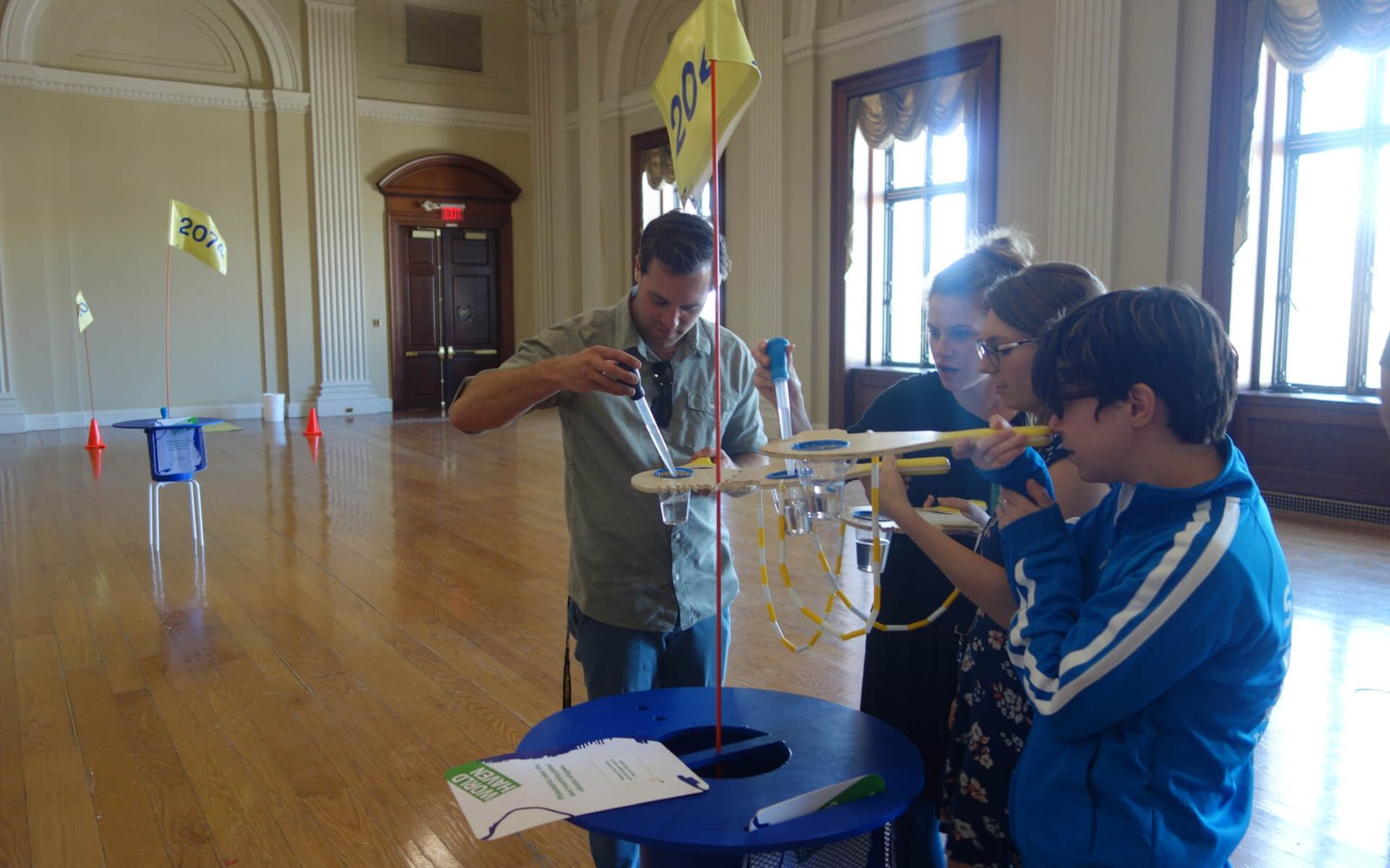 4 people with neighborhood paddles taking on sea level rise by filling their cups with a large dropper