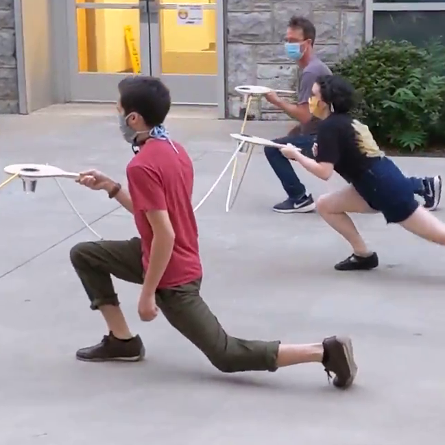 3 people kneel while holding long dash paddles distanced by 6 foot connectors