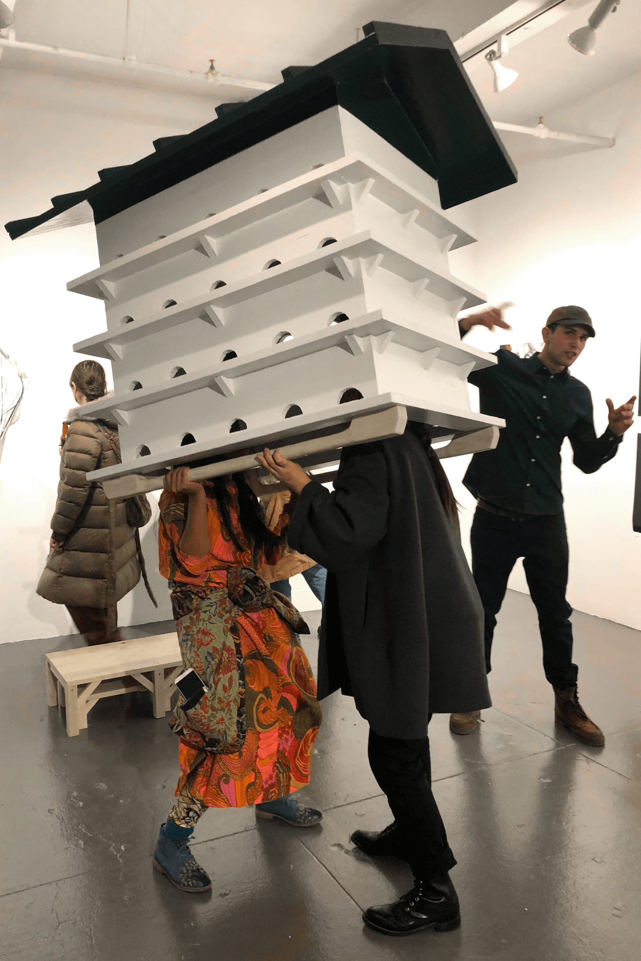 Two people play Martin Dance. They are standing, facing each other about shoulder width apart, with bent legs. Their heads disappear inside a large white purple martin hotel with a green roof.