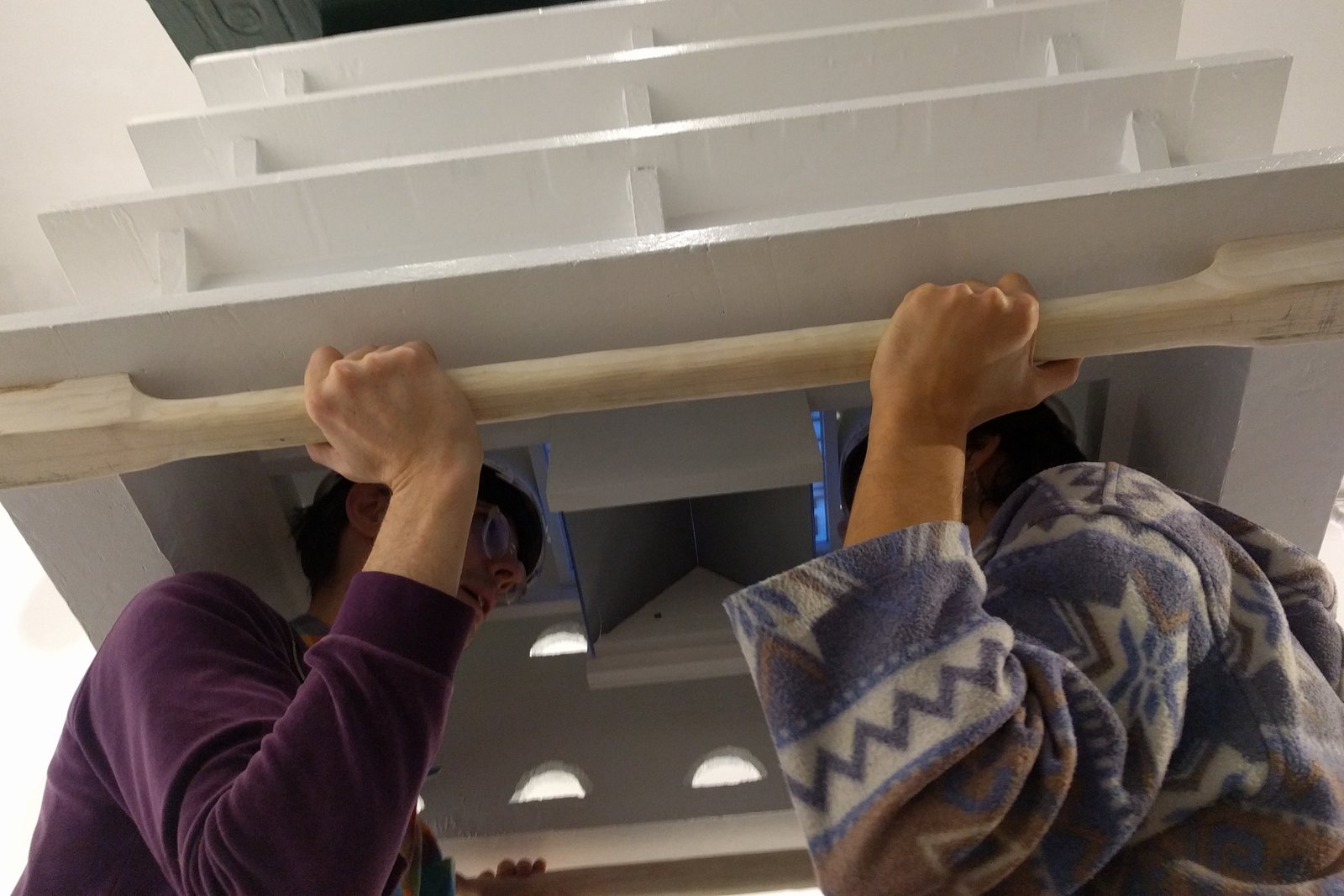 Looking up from just below elbow height, two people's heads are inside a giant bird house. They are each looking at a mirror that reflects a tiny labyrinth they are navigating by moving their bodies.