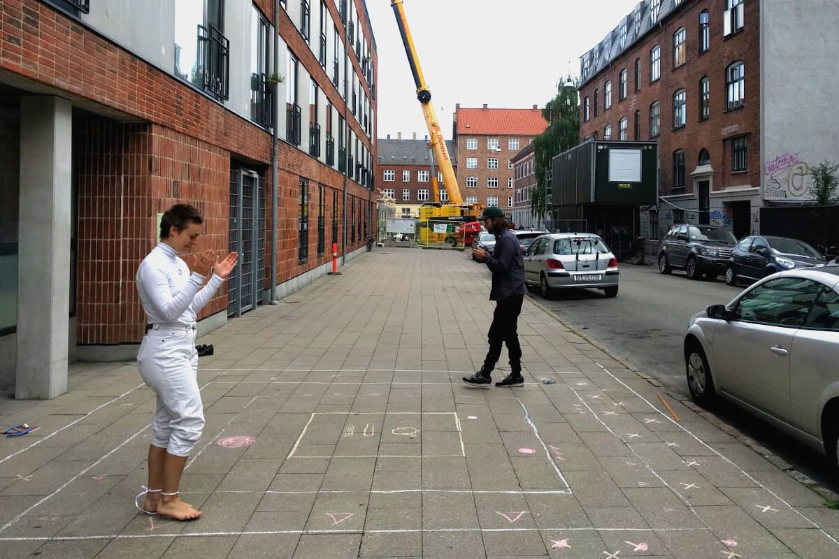 a 5 x 5 meter track drawn in chalk on the sidewalk. Two people are walking in the tracks and clapping when they step on shapes drawn in the tracks